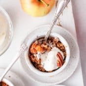 Single ramekin with air fryer apple crisp with whipped cream and pecans surrounded by an apple and partial views of 2 other ramekins of apple crisp.