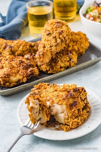 A piece of Air Fryer Cornflake Fried Chicken on a small white plate with other pieces of chicken on a small blue platter with 2 glasses of beer in the background with a blue napkin behind that.
