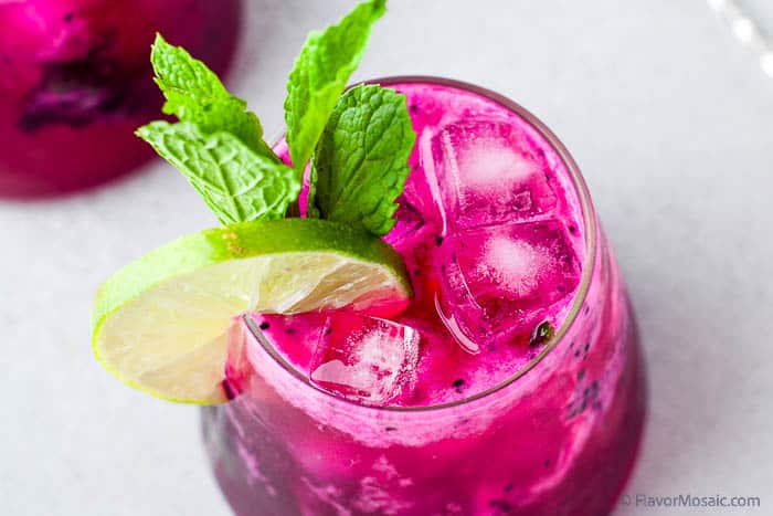 Horizontal photo of a single cocktail glass of dragon fruit mojito on ice with a lime slice and mint leaves. The glass is sitting on a light gray background. A partial view of a second glass of dragon fruit mojito is in the upper left.
