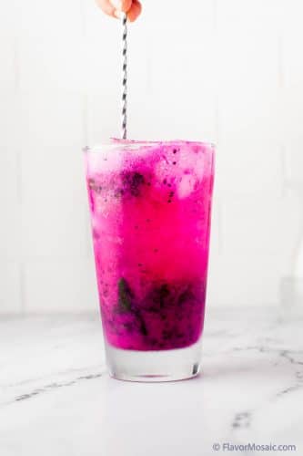 Side view of stirring a high ball glass with a pink dragon fruit mojito with a cocktail stirrer with a white background.