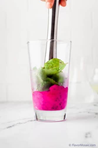 Side view of smashing the mint leaves and dragon fruit in a high ball glass with a muddler.