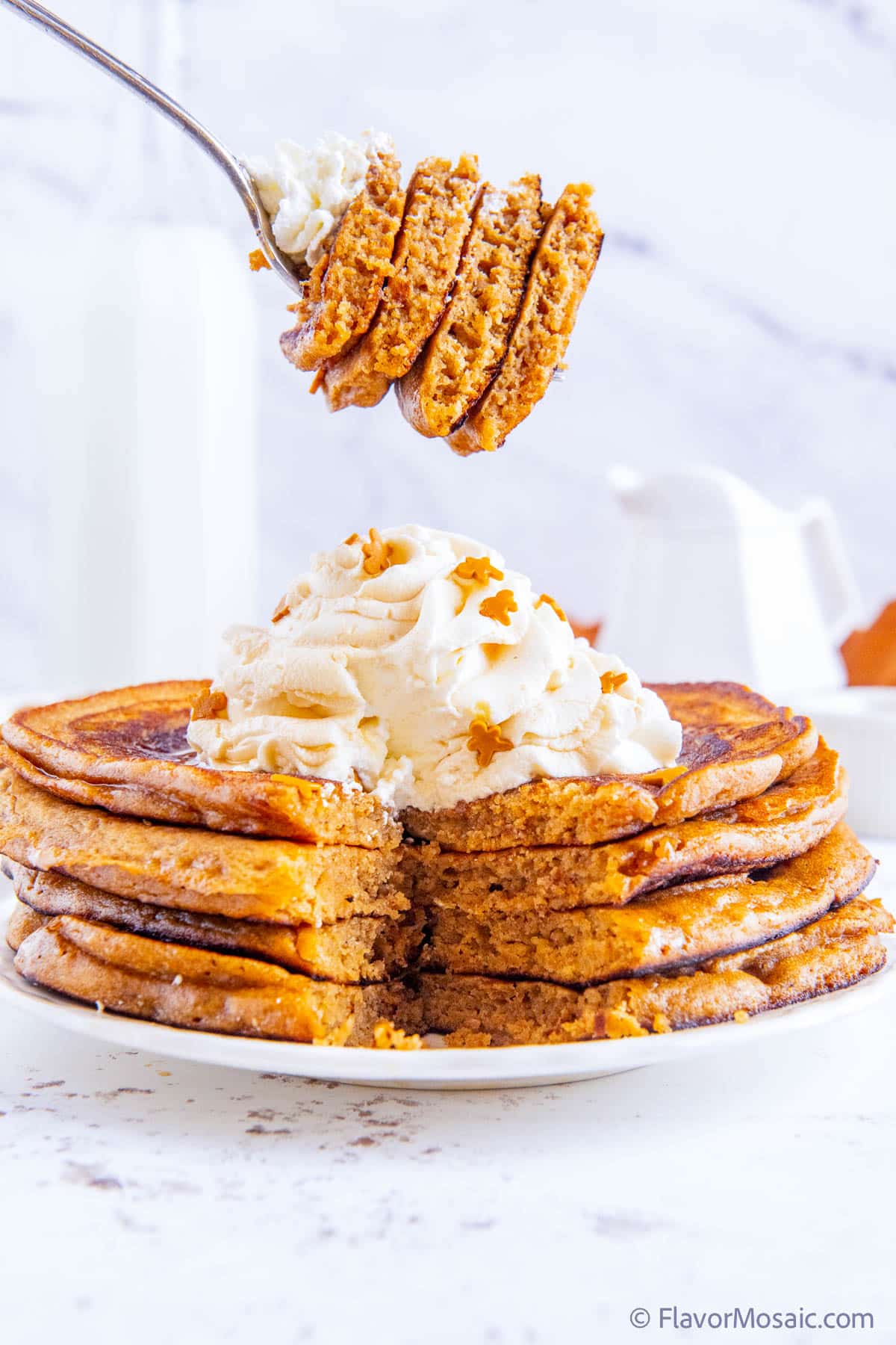 A fork with a bite of 4 gingerbread pancakes with whipped cream being held in the air, against white marble background, and above a stack of 4 gingerbread pancakes, topped with whipped cream, with a single bite cut from all 4 pancakes.