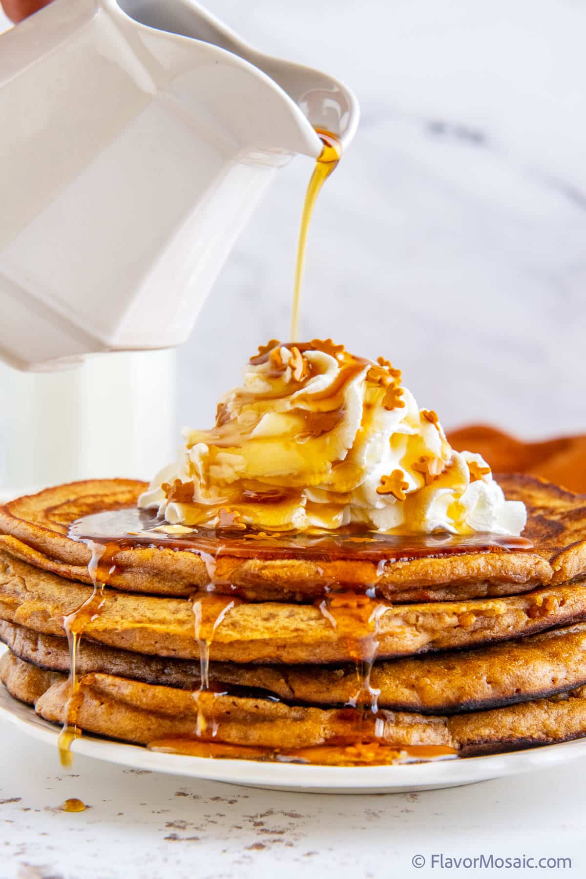 White pitcher pouring maple syrup over a stack of 4 gingerbread pancakes, sitting on a white plate against a white marble background, topped with whipped cream, and maple syrup drizzling over the sides of the pancakes.