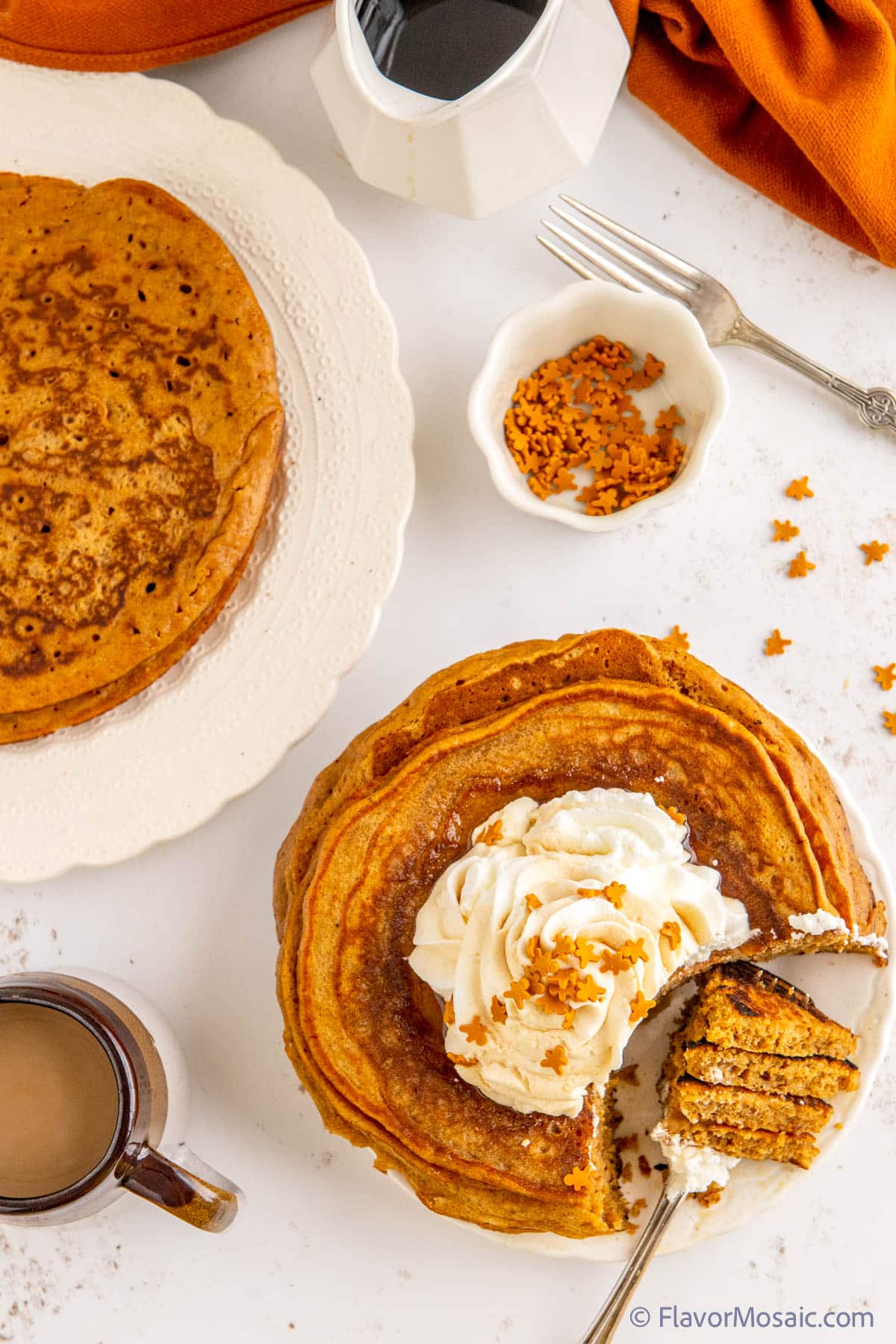 An overhead photo of a white plate with a stack of gingerbread pancakes with whipped cream with tiny gingerbread man edible decorations, with a fork with a bite of 4 pancakes with whipped cream on the fork sitting on the plate. In the upper left is a partial view of a white plate with another stack of pancakes, In the upper right is a rust orange napkin with a small white pitcher with maple syrup, a fork, a small white container of tiny gingerbread man decorations, all on a white granite background.