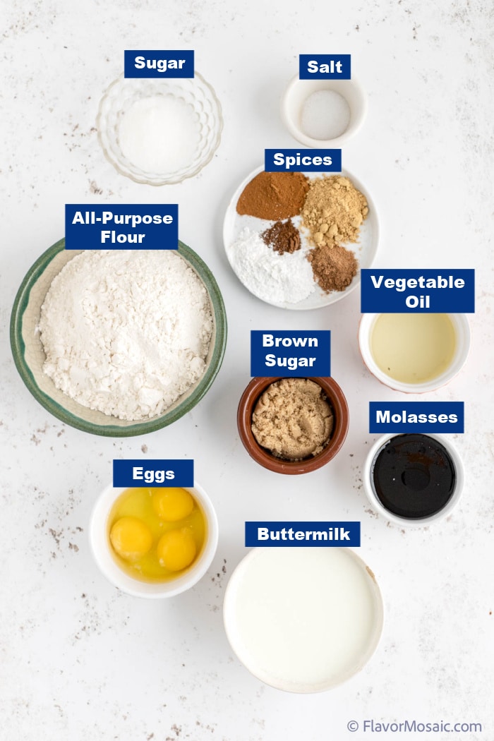 Overhead view of ingredients for gingerbread pancakes, each in an individual bowl or plate, and labeled with a dark blue label with white text, on a white granite background.