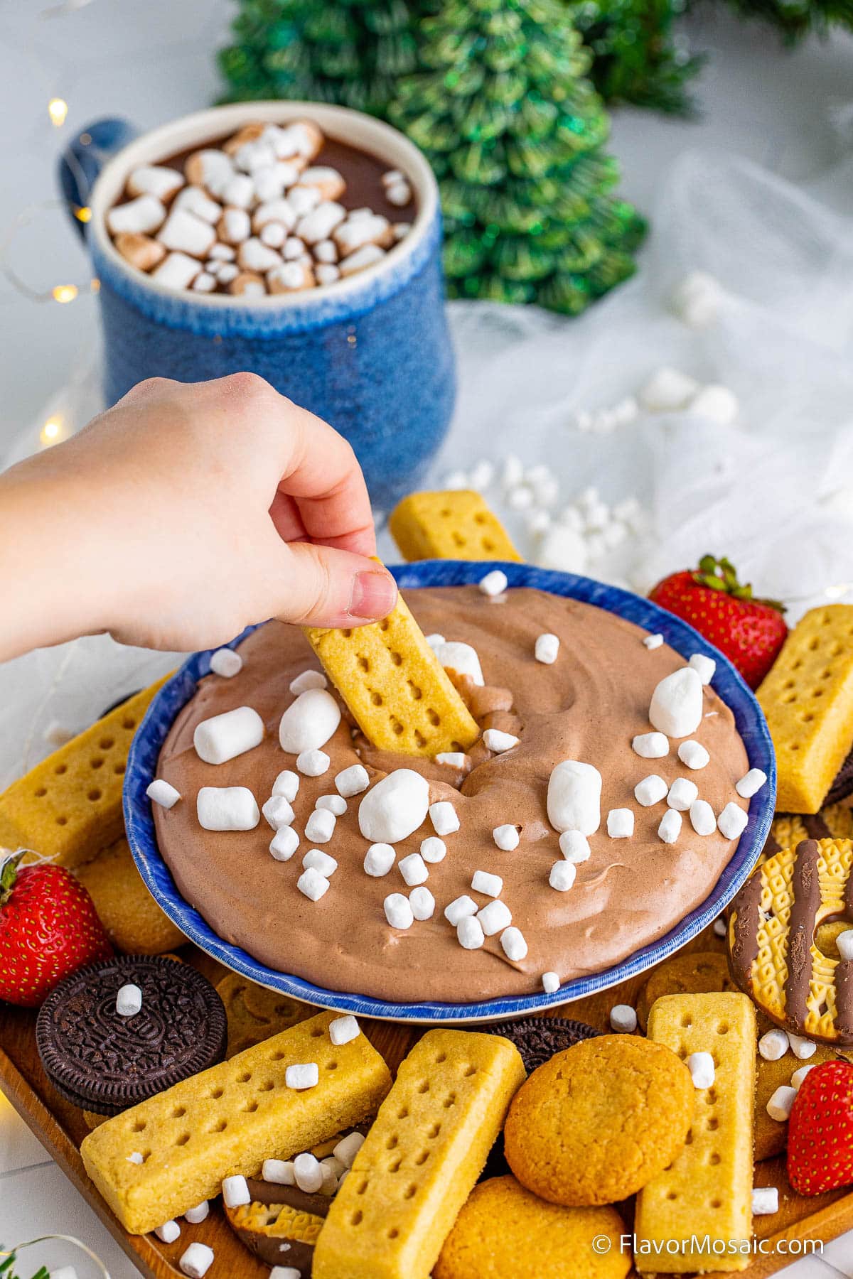 A vertical photo with a view of someone dipping a cookie into a  chocolate dip in a blue bowl topped with mini marshmallows and surrounded by a wood plate of cookies and strawberries.