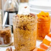 Vertical photo of a side view of a mason jar with pumpkin pie oats with pecans and whipped cream. With and orange and white checkered napkin and spoon on the bottom right, a glass jar of pumpkin puree in back, with a stack of white bowls behind it, a bottle of vanilla in the upper left background, a small glass jar of brown sugar, brown sugar, pecans, and oats scattered about on the white-gray counter, with a small wooden spoon with ground flax seed in the bottom left.