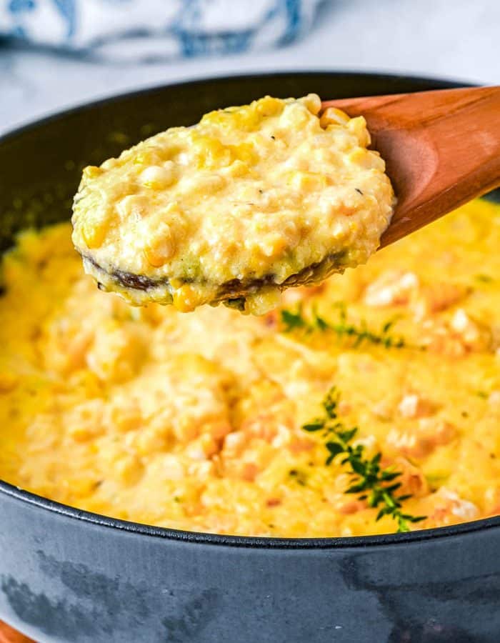 A large wooden spoon with a spoonful of creamed corn held above a large black pan with creamed corn and garnished with fresh thyme.