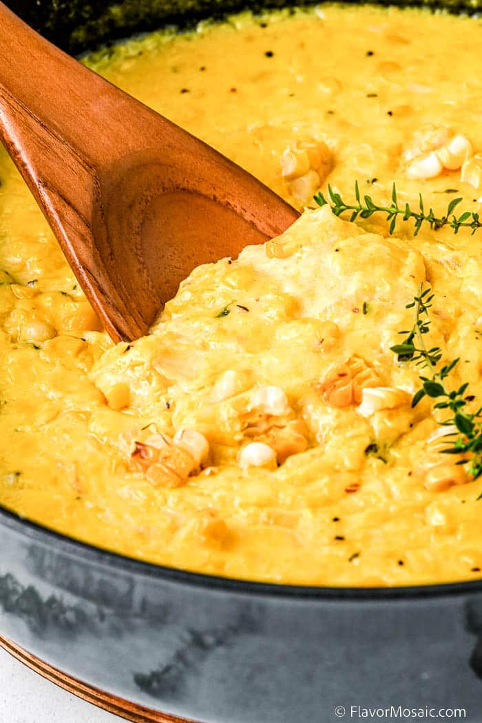 Close up of creamed corn in a skillet garnished with fresh thyme and with corn kernels cut from the cob and a wooden spoon getting ready to get a spoonful of the corn.