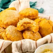 Side view of a towel covered bowl with at least half a dozen sweet potato biscuits with one biscuit sitting on top with a bite taken out of it and the top covered with honey butter. In the back is a green napkin.