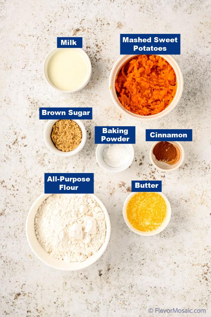 An overhead view of individual bowls, each containing a single ingredient for the sweet potato biscuits recipe. All sitting on an off-white concrete looking counter. Each ingredient is labeled with a navy blue rectangular label with white text.