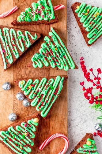 Overhead view of triangle-shaped Christmas Tree Brownies with green frosting and sprinkles sitting on a wood cutting board with small silver balls, with a white/gray countertop in the background and small brightly colored red berries on the right.