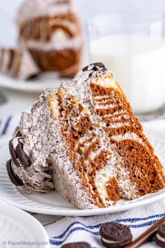 A large slice of zebra cake, with cookies and cream frosting, on a white dessert plate, on a white napkin with blue strips with mini Oreo cookies in front and a glass of milk in back and another slice of cake in the back.