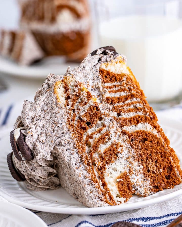 A large slice of zebra cake, with cookies and cream frosting, on a white dessert plate, on a white napkin with blue strips with mini Oreo cookies in front and a glass of milk in back and another slice of cake in the back.