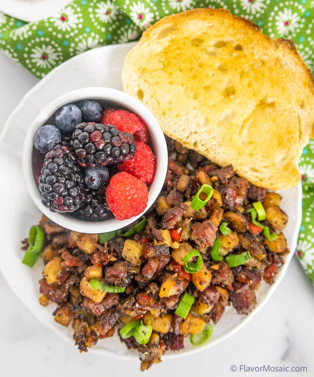 Overhead view of corned beef hash on a white plate with a small white bowl of blackberries and raspberries with a piece of toast next to the hash on the plate with a green napkin with white flowers in the upper right background.