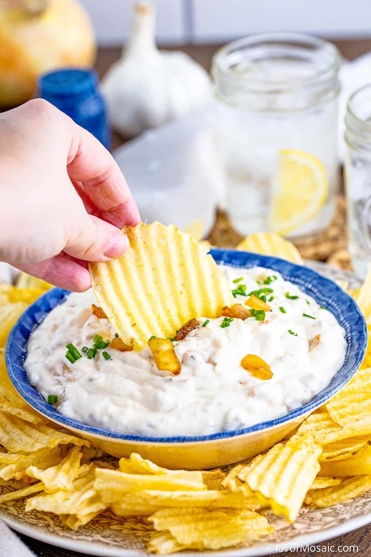 A wavy potato chip is being dipped into a creamy homemade French Onion Dip, with chives and caramelized onions.