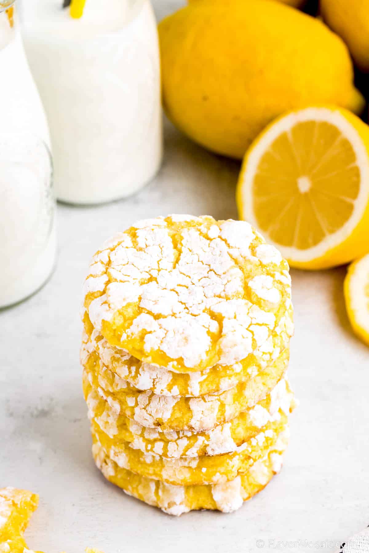 A single vertical stack of Lemon Cool Whip Cookies on a white background with 2 glasses of milk in the upper left background and whole lemons with one lemon cut in half in the upper right background.