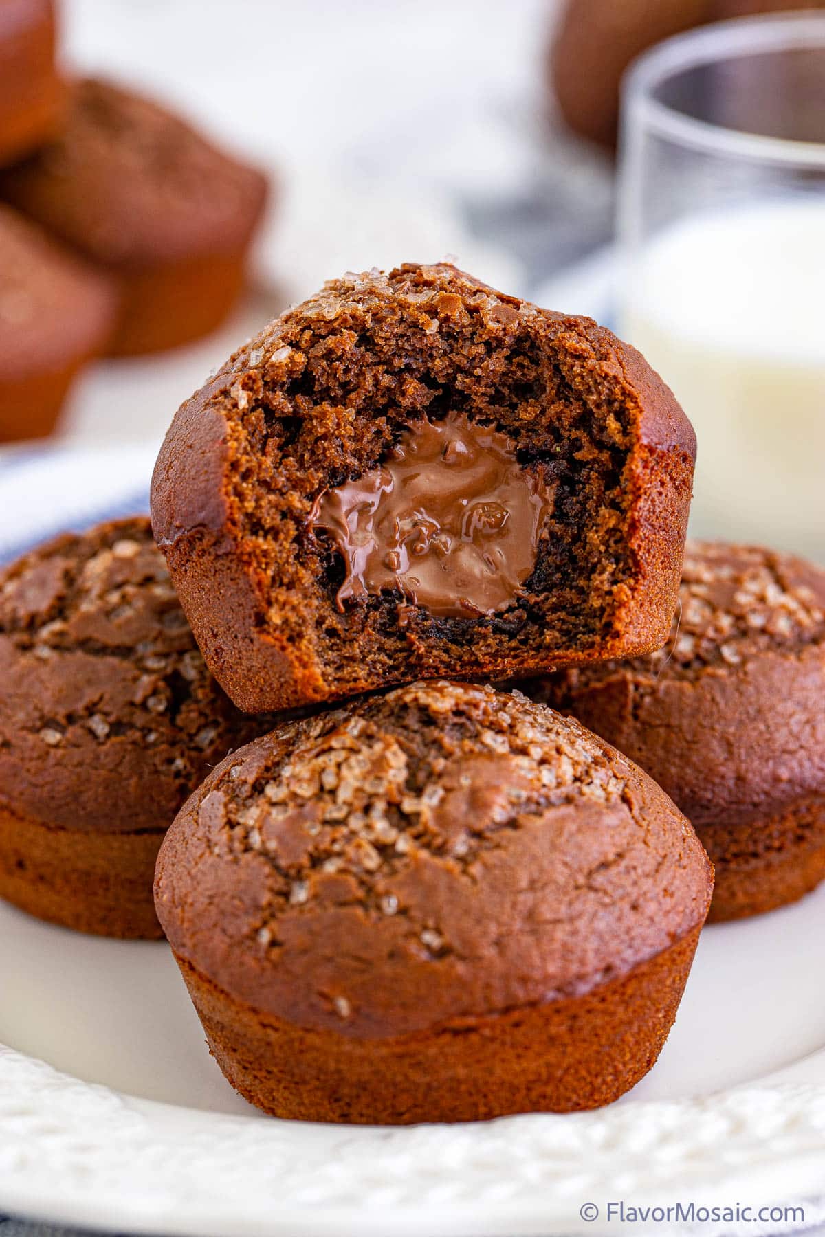 A side view close up of a Nutella Muffin with a bite taken out of it showing the creamy Nutella filling, sitting on top of 3 other Nutella Muffins on a bite plate. In the background is a partial blurred view of a glass of milk and 2 other plates of Nutella Muffins.