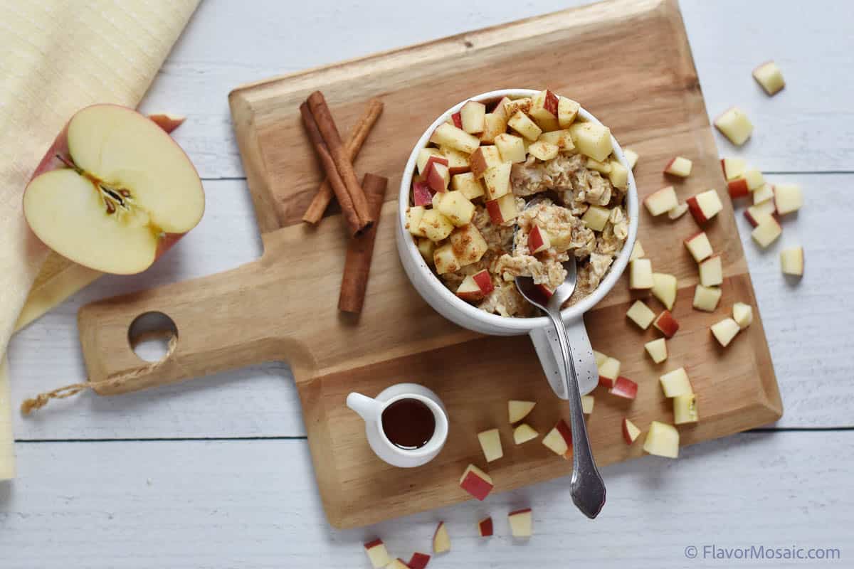 Overhead view of Apple Oatmeal Custard in a mug topped with a chopped red apple sitting on a cutting board with with 2 cinnamon sticks. A half an apple, sliced side up is on the left with a cream colored napkin and chopped apples scattered around on the cutting board.