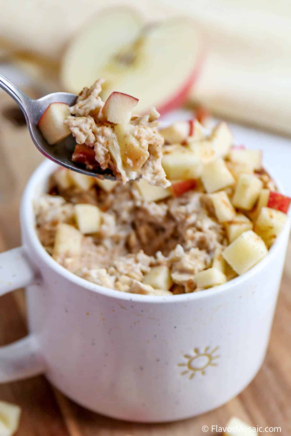 A spoonful of Apple Oatmeal Custard  held over a mug of the oatmeal topped with a chopped red apple.