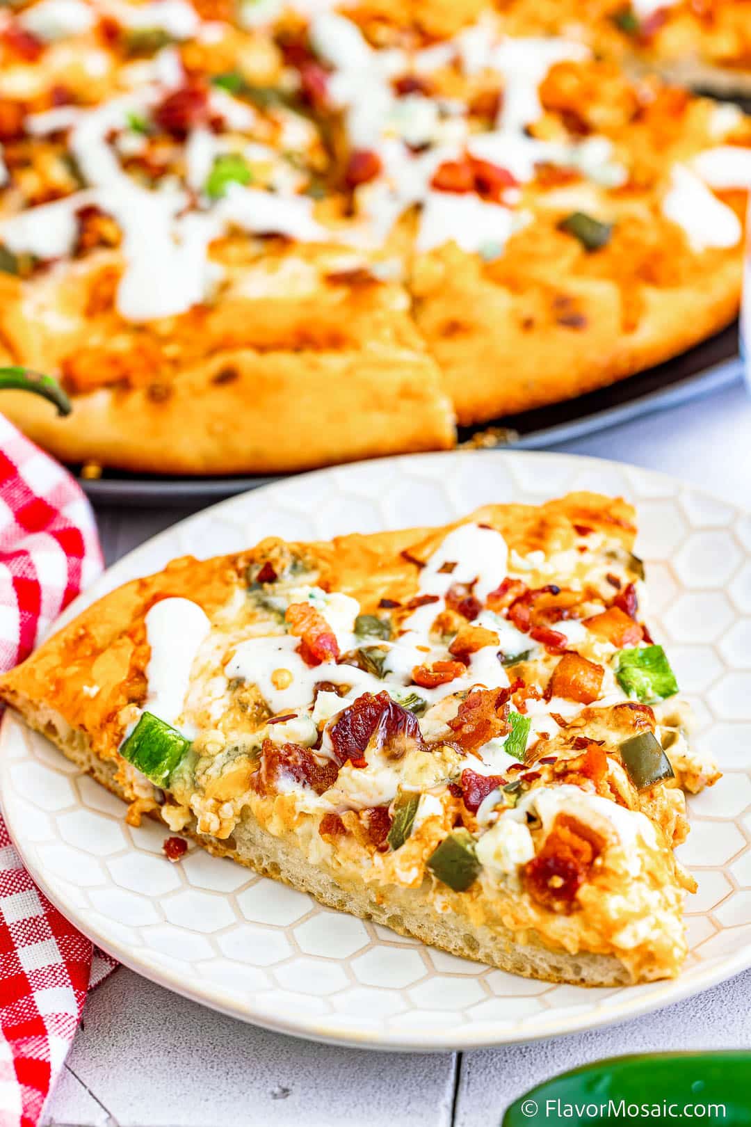A slice of Buffalo Chicken Jalapeno Popper Pizza with a red and white checkered napkin on the left and a partial blurred view of the rest of the pizza on a pizza pan in the background at the top of the photo.