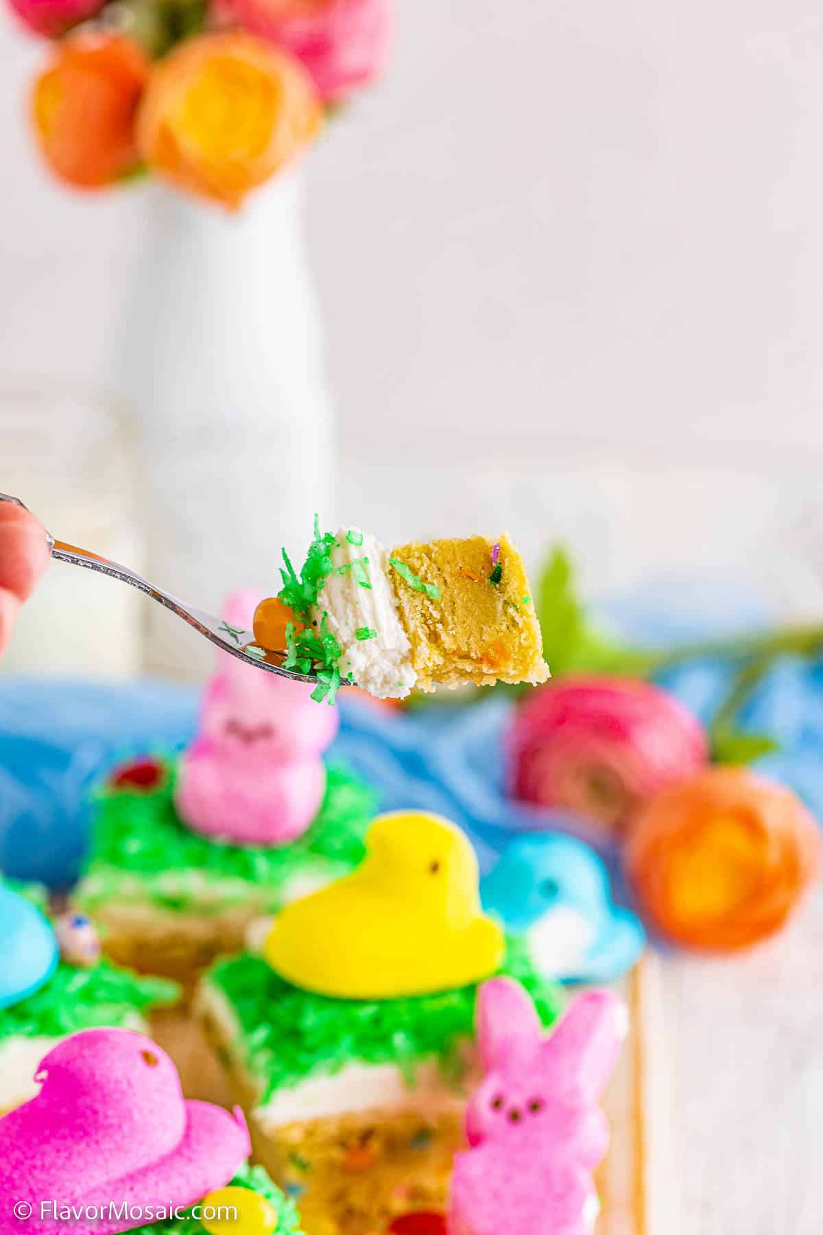 A bite of the Easter sugar cookie with cream cheese frosting, green coconut "grass", and an orange jelly bean sitting on a fork above the other Easter Sugar Cookie Bars with colorful Easter Peeps in the background.