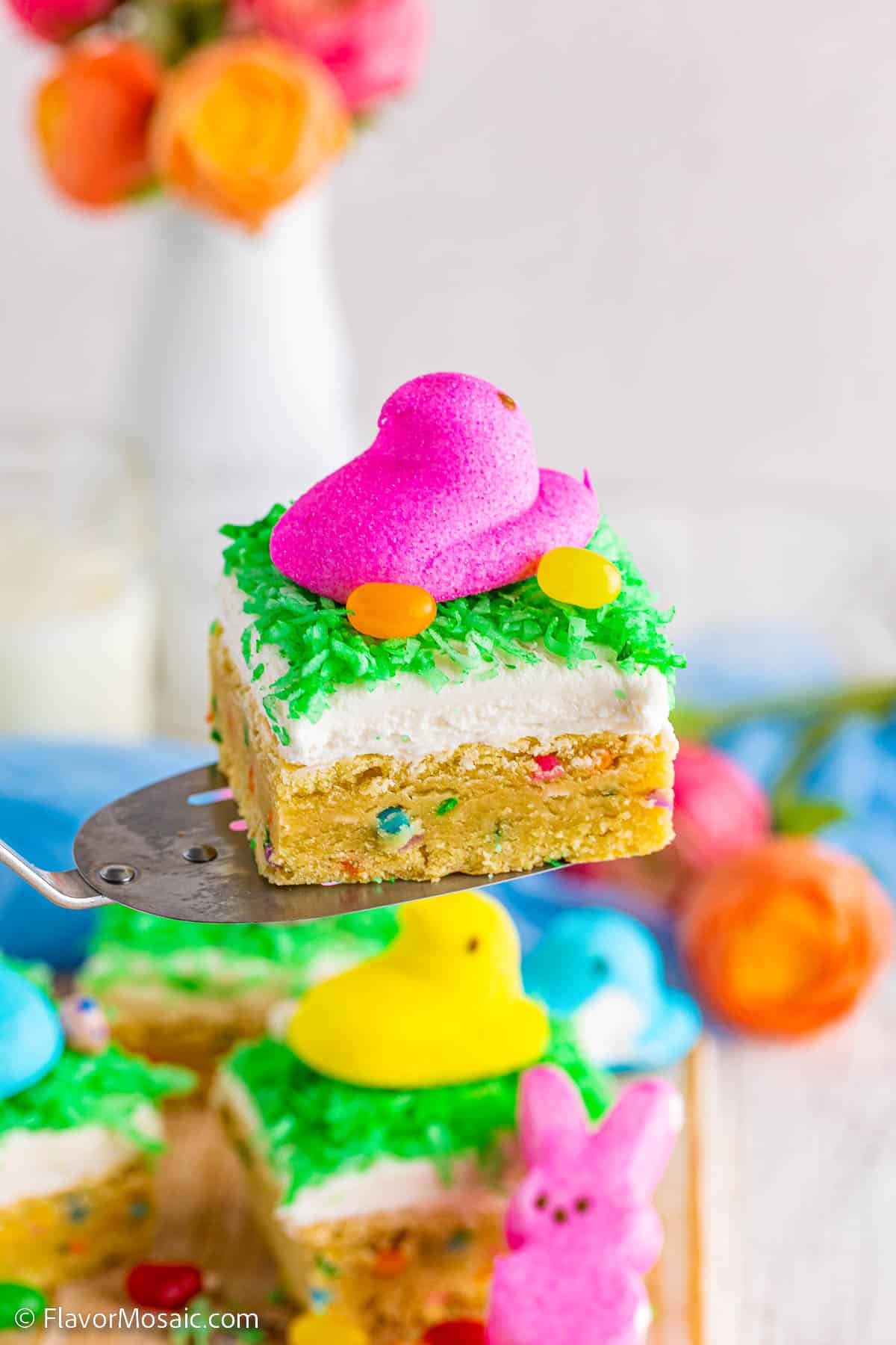 Side view of Easter Sugar Cookie Bar topped with Pink Easter Peep, 2 yellow jelly beans, and green colored shredded coconut held on a spatula above other Peep covered Easter Sugar Cookie Bars.
