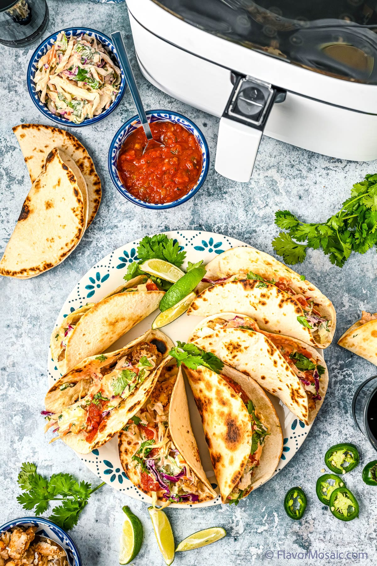 Overhead view of a blue plate with blue decorative edges with 6 air fryer fish tacos on a blue and white cement like background, surrounded by sprigs of cilantro, slimed limes, sliced jalapenos, flour tortillas, a small bowl of red salsa, another small bowl of cilantro lime slaw and a partial view of an air fryer.