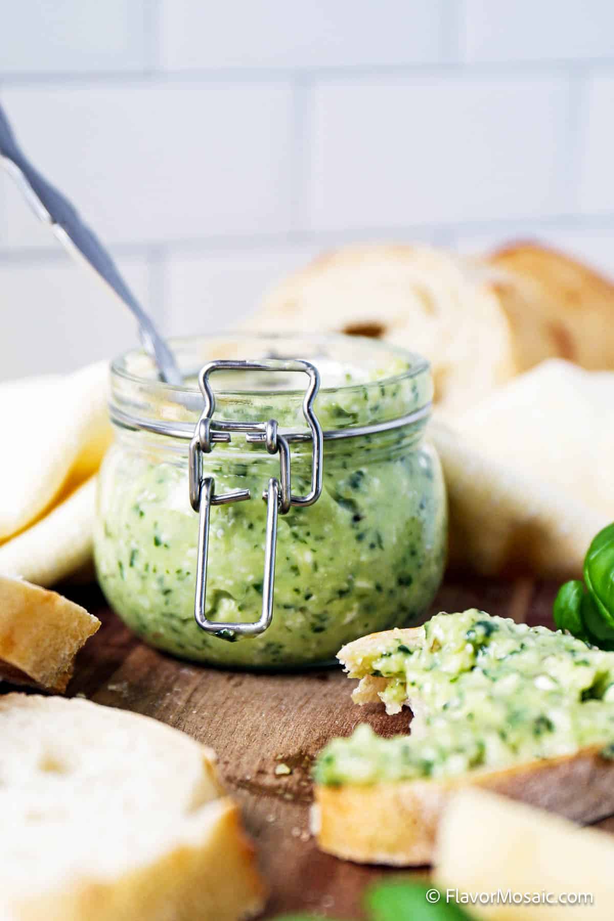 Side view of basil pesto butter in a small mason jar, sitting on a wood cutting board, surrounded by small slices of white French bread. On the bottom right is a partial view of a slice of bread, covered with pesto butter.