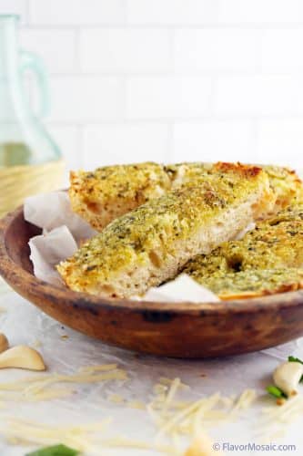 Sliced Pesto Garlic Bread sitting on top of parchment paper inside a wood bowl sitting on more parchment paper surrounded by garlic cloves and shredded parmesan cheese. A glass jar of olive oil in the back.