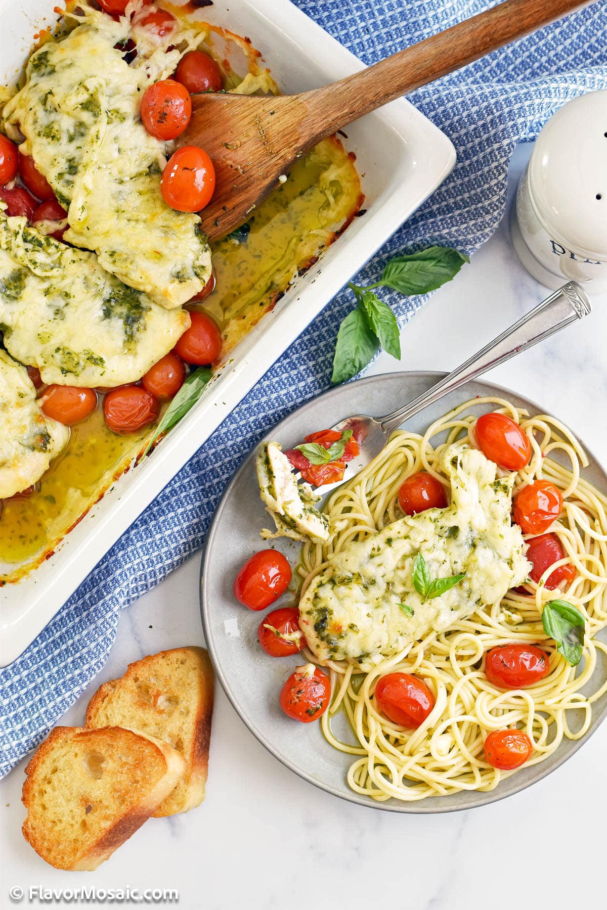 Overhead view of baking dish with cheese covered pesto chicken in the upper left corner, with a plate of a serving of pesto chicken with spaghetti and cherry tomatoes on a blue gray plate on a white background. 2 pieces of toast are on the bottom left.