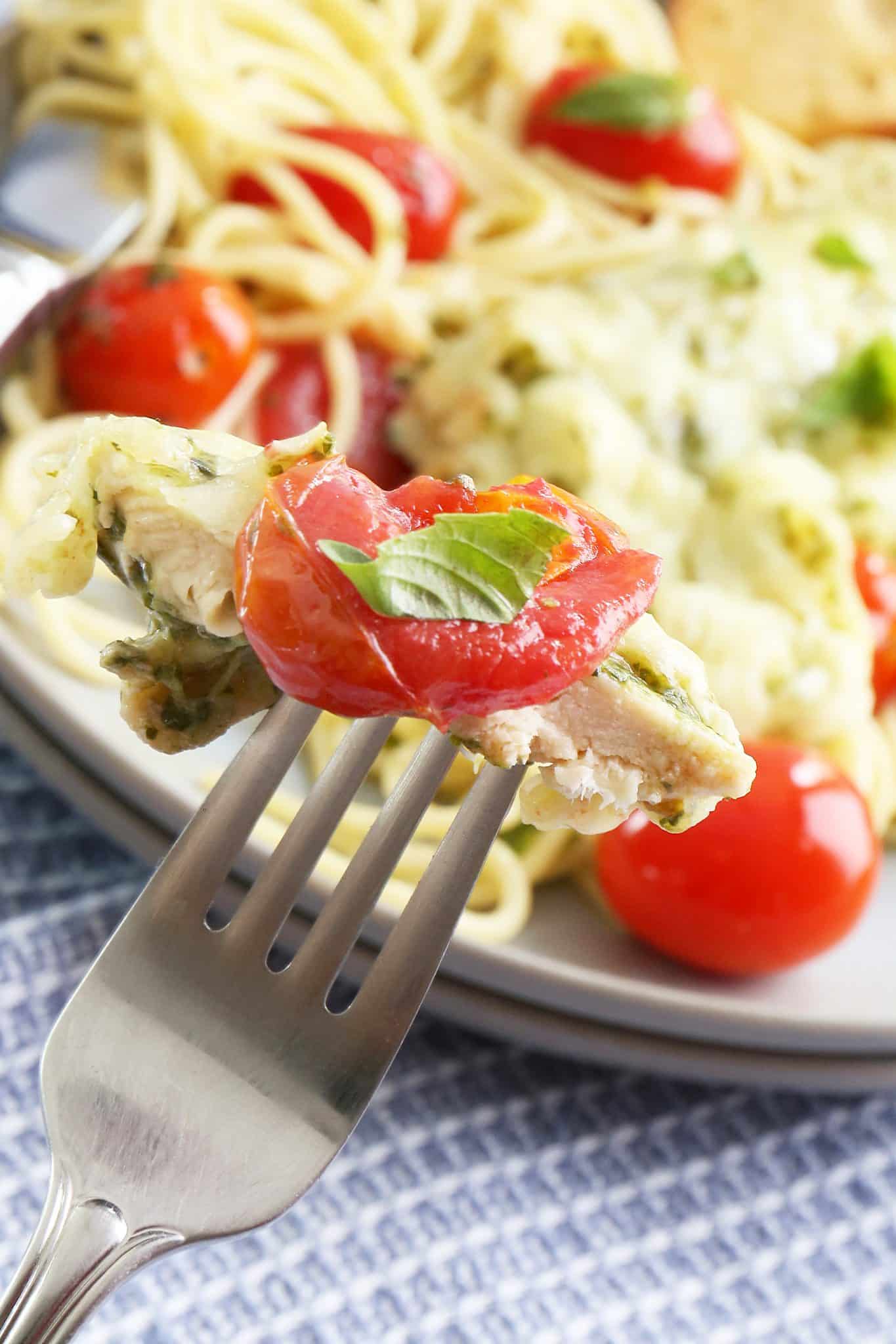A close up of a bite of pesto chicken with a cherry tomato and small piece of basil on a fork held above the rest of the plate of pesto chicken in the background.