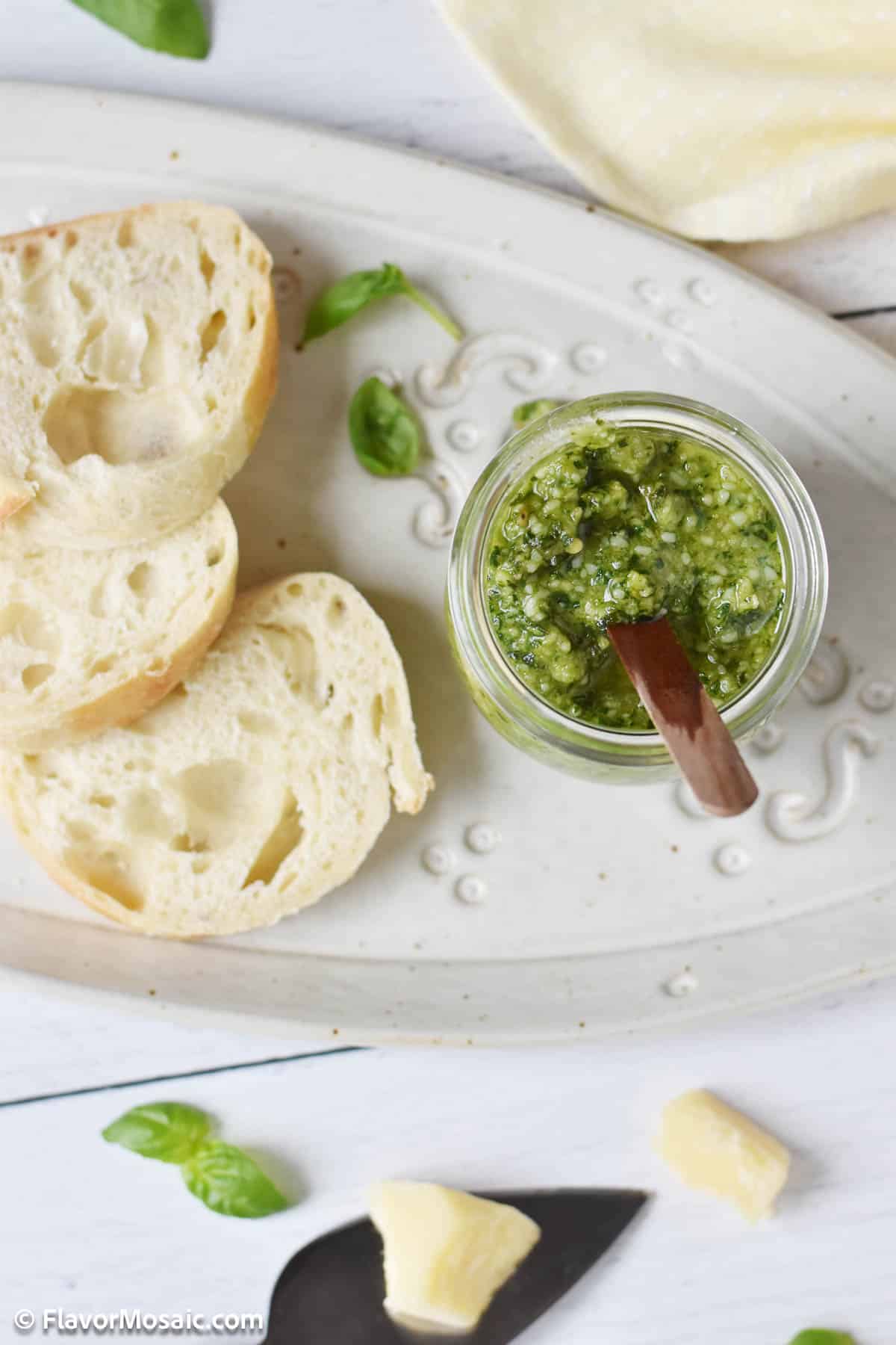 Overhead photo of a jar of pesto with a wooden spoon sticking out of it, sitting on a white plate next to small white pieces of bread. Surrounded by very small basil leaves and chunks of parmesan cheese.