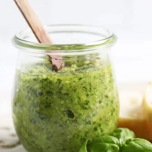 Side view of a clear glass jar of pesto with a wooden spoon in it. Next to the jar of pesto is fresh basil leaves and some slices of bread.