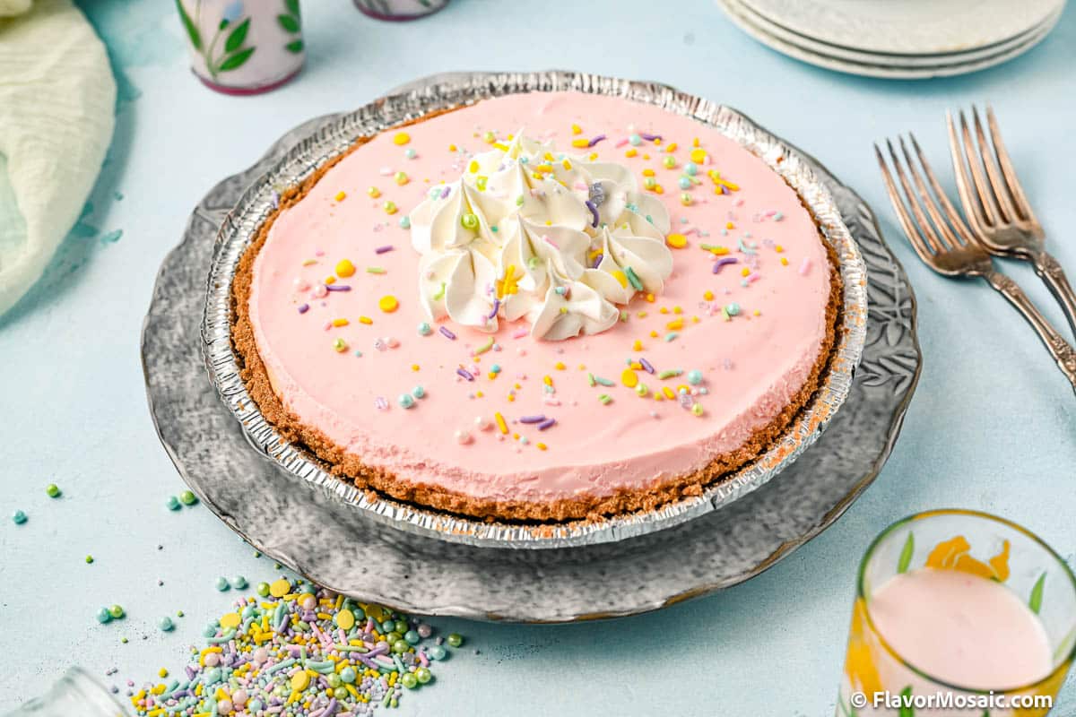 A view of the whole rainbow no bake cream cheese pie on a meetal plate with 2 metal forks on the right, a glass of milk on the bottom right, multi-colored pastel sprinkles on a light blue background on the lower left, and a napkin and partial views of glasses of milk in the upper left.