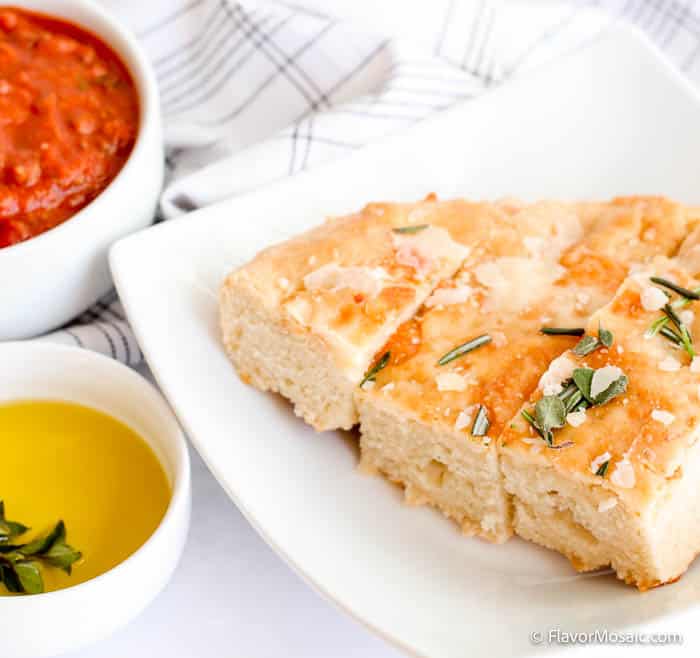An arched white plate with 3 slices of Focaccia bread topped with shaved parmesan and rosemary, next to a white bowl of marinara sauce and a second white bowl of olive oil, with a napkin with gray lines on it in the background.
