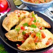 4 Breakfast Empanadas topped with pico de gallo on a black plate with a blue bowl of red salsa in the upper right corner, a tomato, limes and green onion in the upper left, and a white napkin with a wide pink strip on the bottom right.