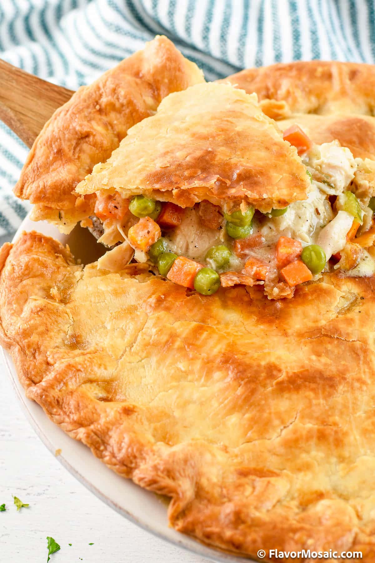 One slice of Chicken Pot Pie Casserole, with its flaky crust, and vegetables in a creamy sauce, is being lifted out of the whole chicken pot pie.