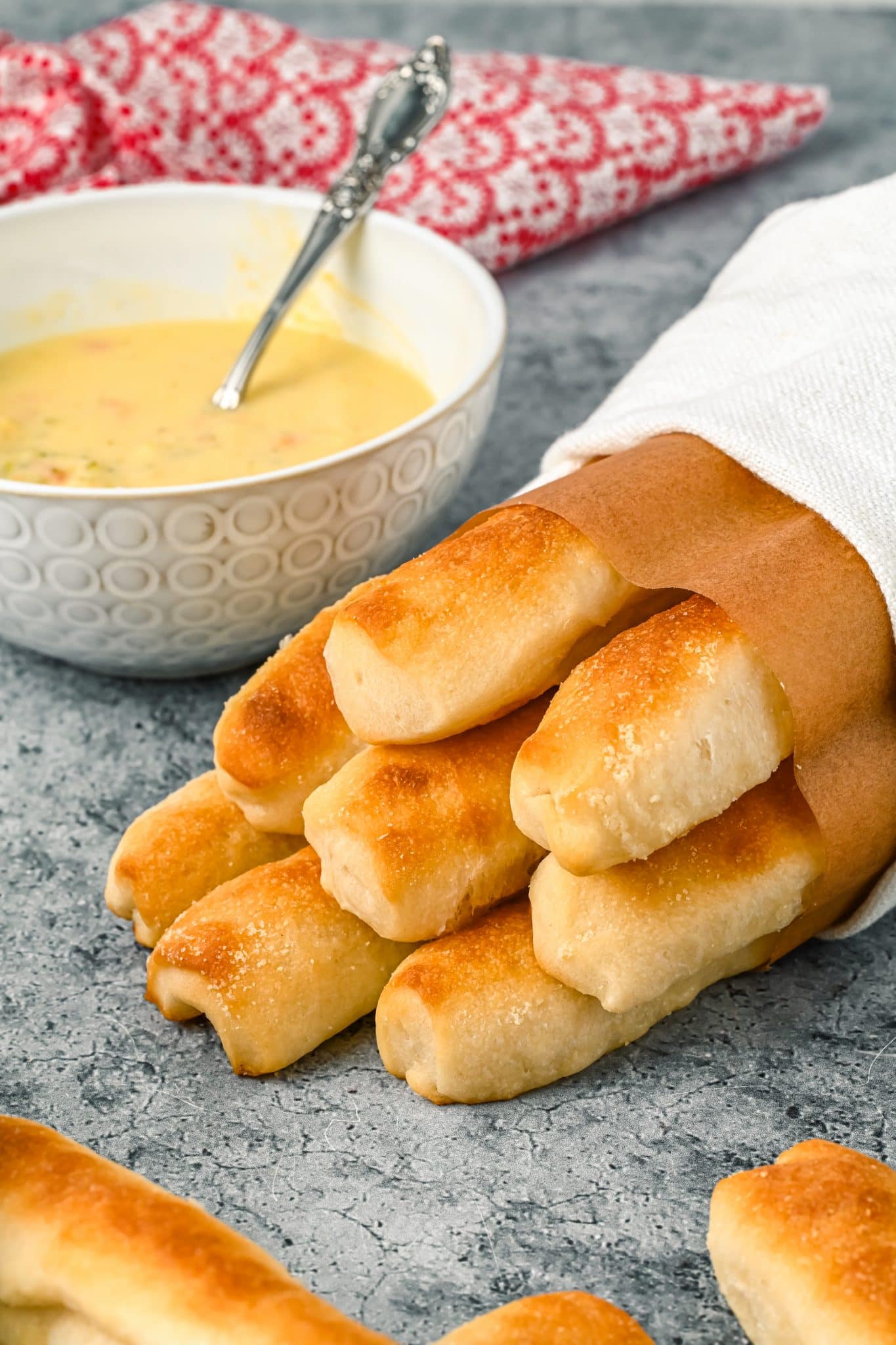 Copycat Olive Garden Breadsticks which are Italian Breadsticks, wrapped in paper, wrapped in a napkin, on a blue/gray background, next to a white bowl of soup.