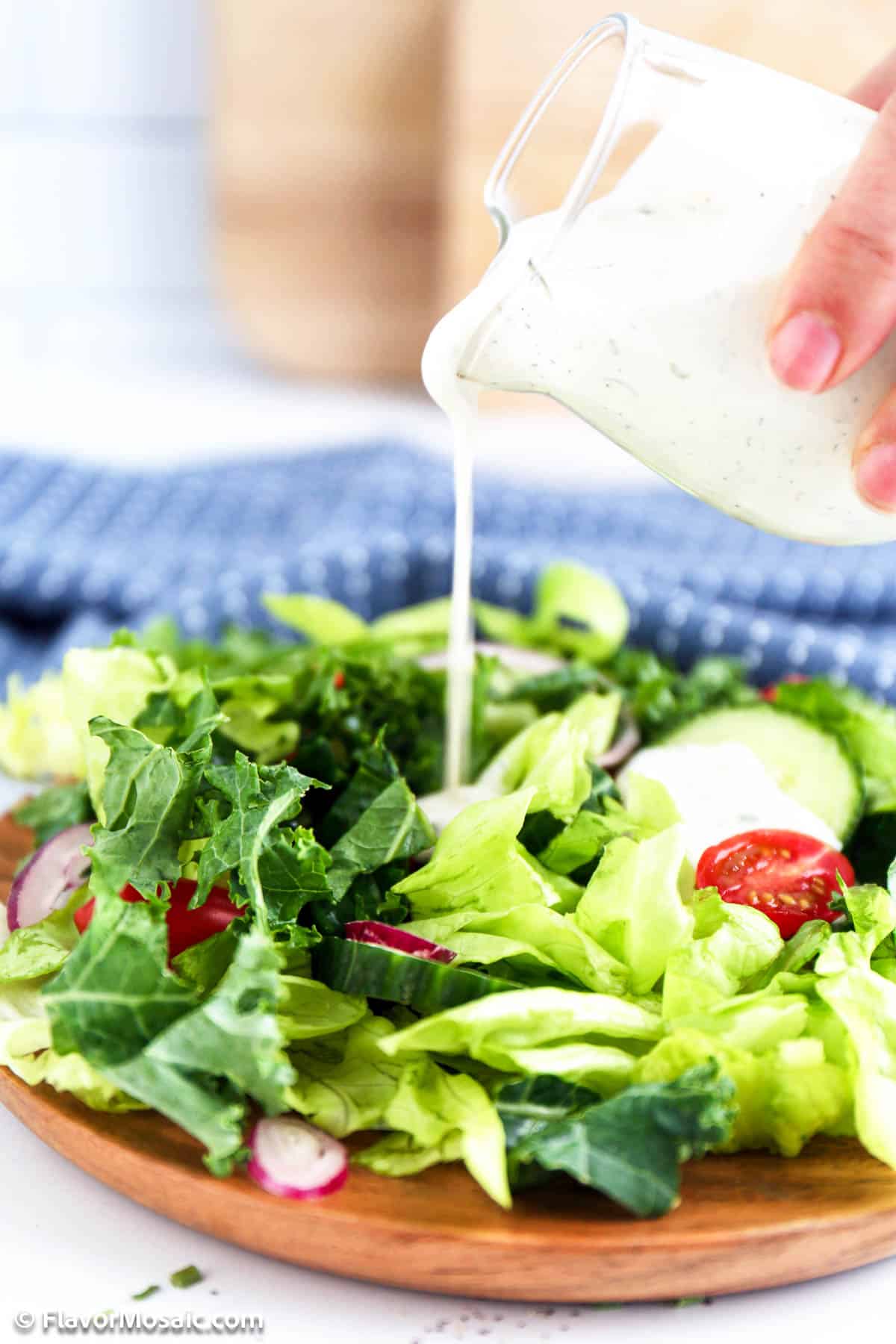 A small glass pitcher filled with ranch dressing being poured over a green leaf salad with cucumbers, tomatoes, radishes, and onions.