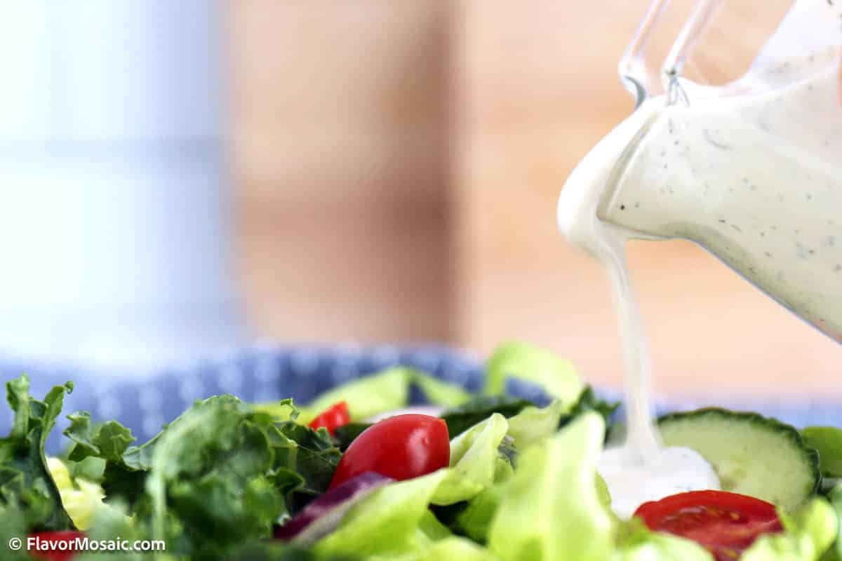 Side view of a glass pitcher being held over a salad pouring ranch dressing over a green salad.