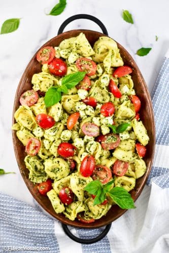 Overhead view of Pesto Tortellini Pasta Salad in a brown dish on a white marble background, surrounded by a blue and white block napkin. The pasta salad is topped with cherry tomato halves and fresh basil leaves.