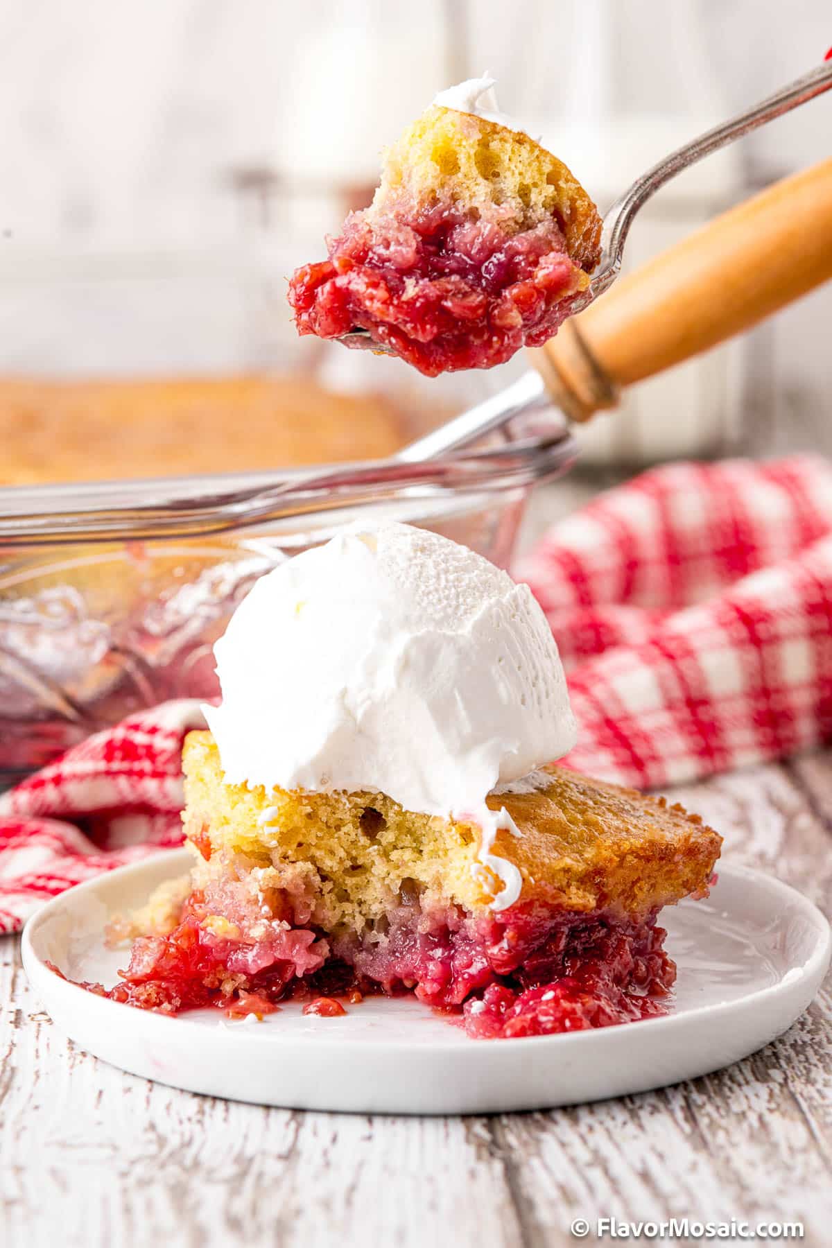 A side view of a white dessert plate with a single serving of raspberry cobbler on it with vanilla ice cream on top that has had a bite taken out of it, and a spoon with a bite of the raspberry cobbler is held above the rest of the serving. In the background is the rest of the cobbler (blurred) in a glass baking dish with a red and white checkered napkin to the right of the dish.