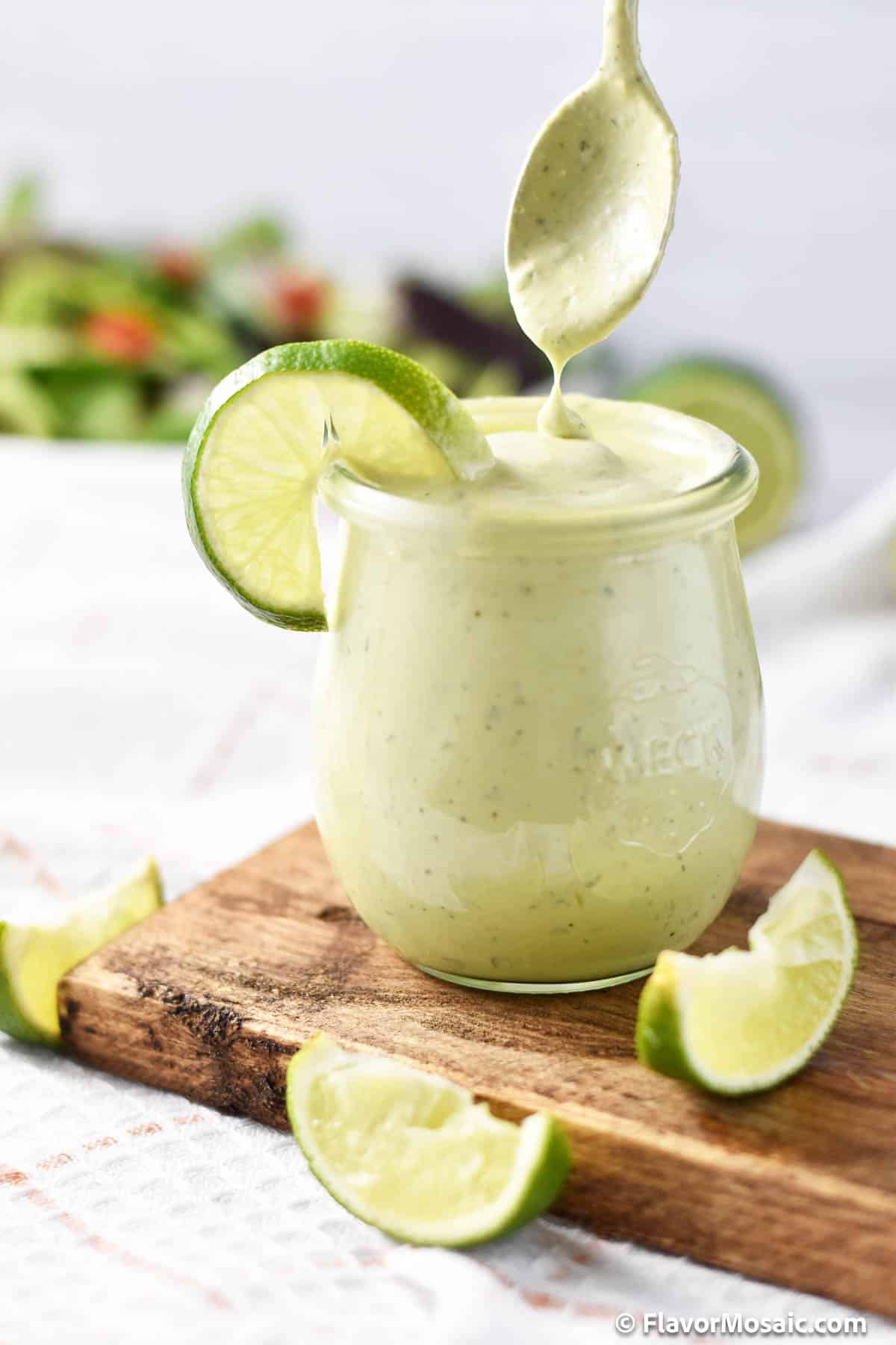 A spoon is held over a glass container of Chick Fil A Avocado Lime Ranch Dressing, sitting on a wood cutting board, in front of a green salad.