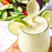 A spoon is held over a glass container of Chick Fil A Avocado Lime Ranch Dressing, sitting on a wood cutting board, in front of a green salad.