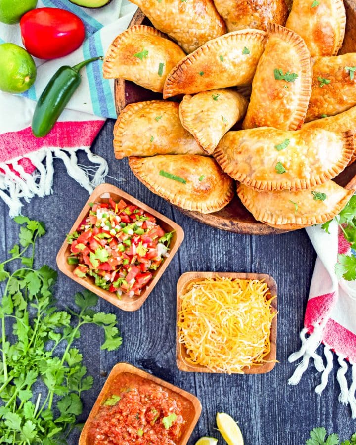 Overhead view of a wood bowl of empanadas with small dishes below it with pico de gallo, cheese, and red salsa. In the upper left corner is a whole jalapeno, tomato, 2 limes, and half an avocado, with the inside facing up.