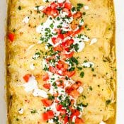 Overhead view of Green Chile Chicken Enchiladas in a white baking dish and garnished with sour cream, chopped tomatoes, and chopped cilantro.