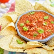 A bowl of red salsa topped with chopped fresh cilantro in a blue bowl surrounded by tortilla chips, with fresh cilantro, tomatoes, garlic cloves, jalapenos, and lime wedges surrounding the bowl.