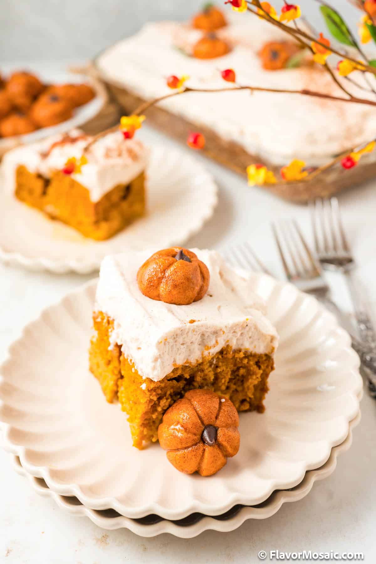 A small white plate with a single serving of Pumpkin Tres Leches Cake topped with a small mini pumpkin with another mini pumpkin on the side. 3 forks next to the plate, with another single serving on a white plate behind that and in the top right is a whole 9x13 cake pan with Pumpkin Tres Leches Cake topped with mini pumpkin and a sprinkle of cinnamon. In the upper left of the photo is a partial view of a white plate with multiple mini pumpkin decorations.
