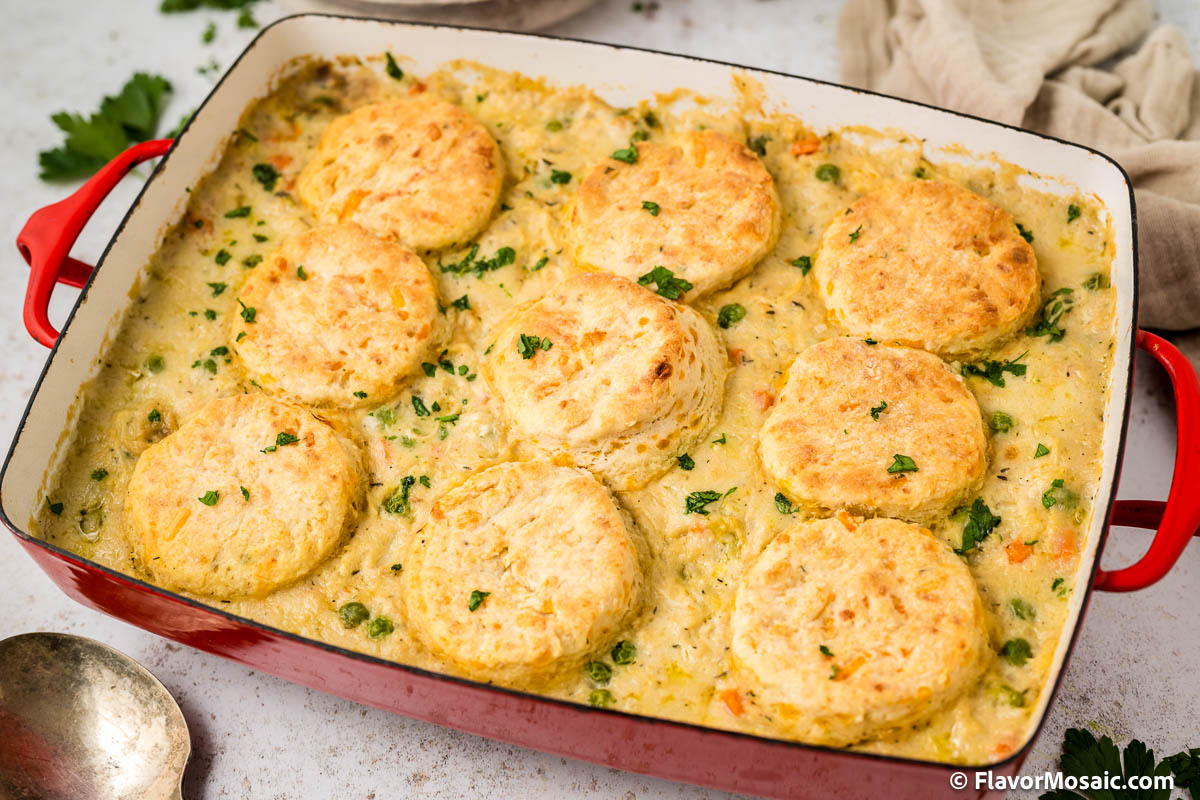 Horizontal photo of an entire red casserole dish with creamed chicken and biscuits casserole with a beige napkin in the upper right and a large spoon in the bottom left with chopped parsley sprinkled on top of the casserole and larger pieces of parsley surrounding the casserole dish in the upper left and bottom right of the casserole.